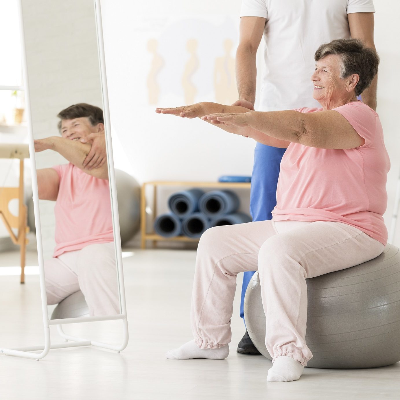 Smiling senior exercising with a physiotherapist on a silver ball at a white gym